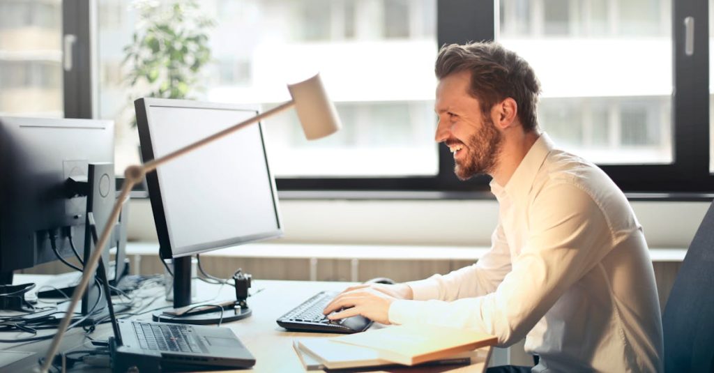 A man smiling while working at an office desk with a computer and natural daylight streaming in through large windows.